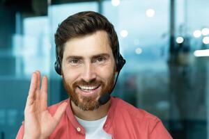 Close-up photo. Portrait of a young man in a headset and red shirt, smiling, working in the office, looking at the camera, gesturing with his hand, explaining. photo