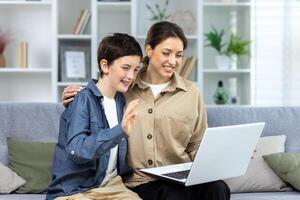 Portrait of family, mother and teenage son sitting on sofa at home and hugging. They are holding a laptop, waving at the camera, greeting each other, talking via call. photo