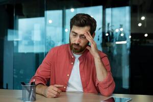 Shocked and upset young man sitting at desk in office, thoughtfully holding head with hand, closed eyes, crying, feeling pain Fired from job, salary reduction, unemployed. photo
