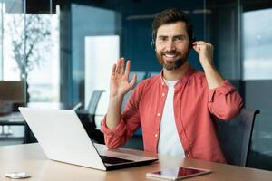 A young man in a headset of a psychologist, psychotherapist, sitting in the office at the table with a laptop and consulting online, conducting a lecture, looking and greeting the camera smiling photo