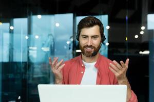 Distance Learning. Portrait of a young man teacher in a headset in a red shirt sits in the office at the table, works on a laptop online, teaches through a call. Explains with his hands, smiles photo