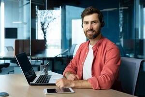 Portrait of mature businessman in red shirt inside office, man with beard and call headset smiling and looking at camera, online customer support worker with laptop. photo