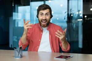 retrato de un joven hombre en un auriculares quien es emocionado hablando a el cámara, ondulación su manos expresivamente. se sienta en el oficina a el mesa, comunica vía llamar, consultas, comentarista. foto