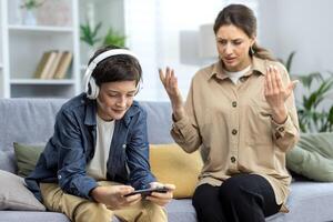 Family conflict quarrel, woman mother quarrels with son, teenage boy in headphones ignores woman and plays games on phone, family at home in living room sitting on sofa. photo