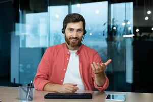 Portrait of a young male blogger in a headset sitting at a desk in the office with a camera and talking via call, recording a podcast, developing his online channel. photo