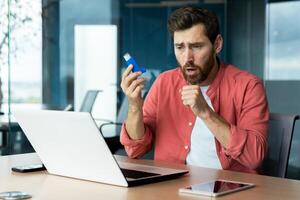 Asthma and respiratory problems in office worker, mature man coughing and using medicine in inhaler to ease breathing at workplace, mature businessman inside office with laptop. photo