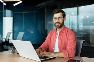 Close-up portrait of mature businessman inside office, man with beard and red shirt smiling and looking at camera, programmer developer coding software at workplace with laptop. photo