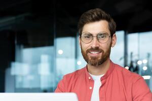 Close-up portrait of mature businessman inside office, man with beard and red shirt smiling and looking at camera, programmer developer coding software at workplace with laptop. photo