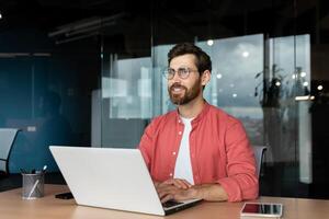 Smiling dreamy businessman inside office at workplace, experienced and successful programmer engineer smiling and looking out window, satisfied with results of achievement at work. photo