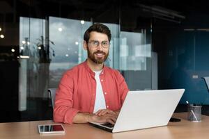 Close-up portrait of mature businessman inside office, man with beard and red shirt smiling and looking at camera, programmer developer coding software at workplace with laptop. photo