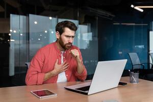 Sick man at workplace, mature worker in red shirt coughing, businessman inside office at work using laptop. photo