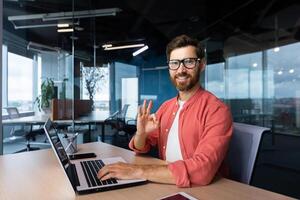 Portrait of a young male programmer, a freelancer who is reading in the office, sitting behind a computer, working on a laptop. He looks at the camera, smiles, shows with his hand ok. photo