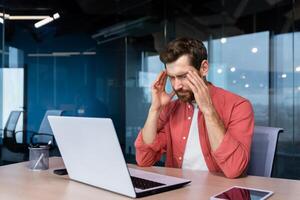Stress and headache at work. A young businessman in a red shirt is sitting at the desk in the office, holding his head, close eyes. He feels bad, worries about problems, a project, overtime, photo