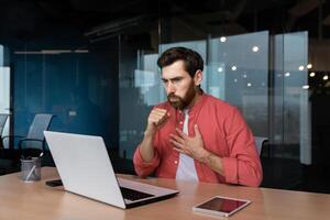 enfermo hombre a lugar de trabajo, maduro trabajador en rojo camisa tos, empresario dentro oficina a trabajo utilizando ordenador portátil. foto