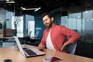 Back pain at the workplace. A young male programmer, freelancer sits in the office at the table and holds his back twisted, stretched, tension pain. Tired from an overtime day. photo