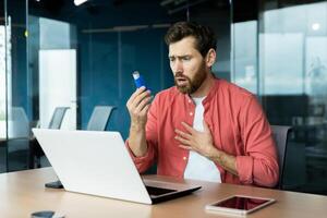 Asthma and respiratory problems in office worker, mature man coughing and using medicine in inhaler to ease breathing at workplace, mature businessman inside office with laptop. photo