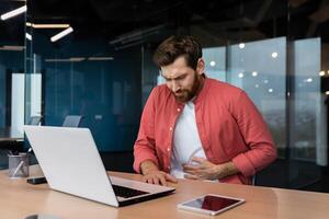 A man in the office at the workplace is sick, he has a severe stomach ache, the businessman got food poisoning from lunch, he is sitting at the table inside the building. photo