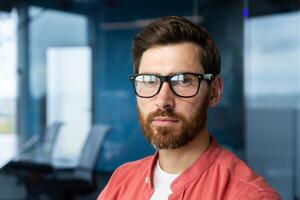 Close-up photo. Portrait of a young professional man, a developer in glasses and a red shirt who looks confidently into the camera. photo