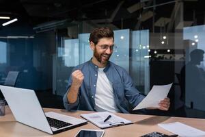 A young man, a businessman, a freelancer sits in the office at the table, holds a document, paper in his hands. He is happy, he points with his hand yes. I received good news, a successful deal, investment, loan. photo