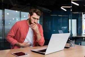 A man in the office at the workplace is sick, he has a severe stomach ache, the businessman got food poisoning from lunch, he is sitting at the table inside the building. photo