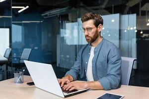 Serious young man freelancer, designer, IT specialist works concentratedly in the office, co-working space. Sitting at a table with a laptop. photo