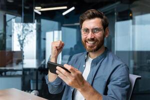 Portrait of man at workplace with smartphone, businessman looking at camera and smiling, using phone for games in free time, worker at workplace inside office, programmer celebrating victory in game. photo
