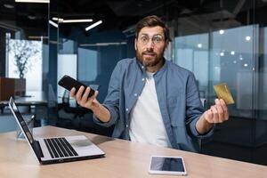 Debt, bankruptcy, foreclosure, fraud. A young man is sitting in the office at the table, holding a phone and a credit card in his hands. He looks worriedly at the camera, spreads his hands. photo