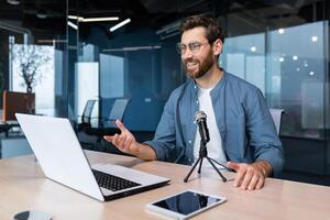 A smiling young man in glasses sits in the office at the table, talks on a call from a laptop into a microphone. writes a blog, podcast, interview, conducts a webinar, business training. photo