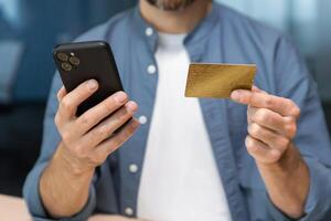 Close-up of man's hands inside the office, businessman in blue shirt and white t-shirt works at workplace, man holds bank credit card and smartphone, makes online purchase in online store. photo