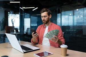 un joven empresario, persona de libre dedicación, banquero se sienta en el oficina a un mesa con un computadora portátil, sostiene efectivo dinero en su manos, usos un teléfono. foto