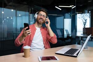 A young male student in headphones studies in the office, campus, at home in front of a laptop, holds a phone in his hand, dances, listens to a lecture. Smiling, resting. photo