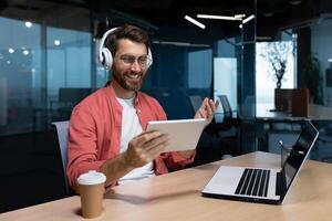 A young businessman freelancer, student, programmer sits in the office at the table with a laptop in headphones, holds and uses a tablet. Talks on a call, listens to music. photo