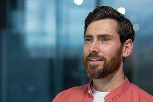 Close-up photo. Portrait of a successful and smiling young man with a beard and wearing a red shirt sitting in the office and confidently looking away. Thinks, dreams, poses. photo