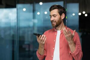 A businessman in a casual shirt near the window is recording an audio message and using an online assistant translator, a man inside the office is satisfied and smiling, using smartphone. photo