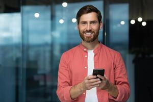 retrato de un joven hombre en un rojo camisa en pie en el oficina, participación un teléfono en su manos, mirando a el cámara con un sonrisa. foto
