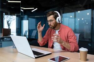 Worried young male freelancer, programmer in headphones, sitting at a table and working on a laptop, talking on a call. Problems in the office. photo
