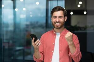 retrato de un joven empresario en pie en el oficina, participación un móvil teléfono y contento demostración un victoria gesto con su mano. concluido un exitoso trato, proyecto, recibido bueno noticias. foto