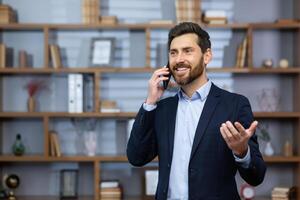 Mature successful boss happy with achievement talking on phone inside office, senior lawyer consulting clients on phone, businessman wearing glasses in business suit, man at workplace standing. photo