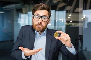 Close-up photo. Confused young business man holds a gold coin, cryptocurrency, bitcoin in his hand. He looks into the camera, points to a coin, points with his hand, asks a question. photo