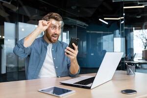 maduro hombre conmocionado por malo Noticias y en línea notificación recibido en teléfono, empresario a trabajo frustrado y gritos a teléfono inteligente pantalla, trabajador a lugar de trabajo sentado con ordenador portátil. foto