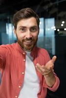 Vertical photo. Close-up photo. Young man freelancer, blogger in red shirt greets and talks to the camera, smiles, waves, records , blog. photo