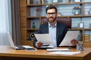 Portrait of successful financier, man with documents in hands smiling and looking at camera, businessman behind paper work sitting at desk inside office, mature boss in glasses and business suit. photo