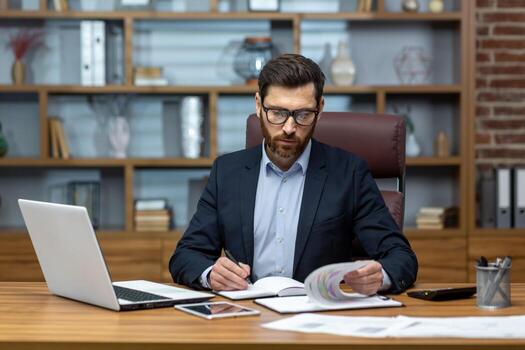 Serious and focused financier accountant on paper work inside office, mature man using calculator and laptop for calculating reports and summarizing accounts, businessman at work. photo