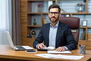 retrato de exitoso empresario detrás papel trabajar, hombre en camisa sonriente y mirando a cámara, financiero jefe dentro oficina utilizando ordenador portátil en trabajo a lugar de trabajo. foto