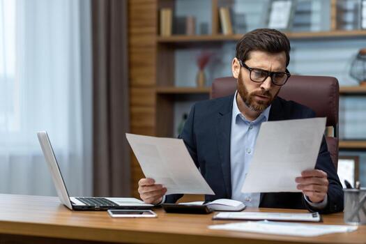 Serious and focused financier accountant on paper work inside office, mature man using calculator and laptop for calculating reports and summarizing accounts, businessman at work . photo