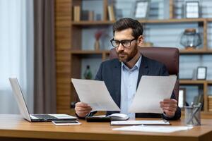 Serious and focused financier accountant on paper work inside office, mature man using calculator and laptop for calculating reports and summarizing accounts, businessman at work. photo