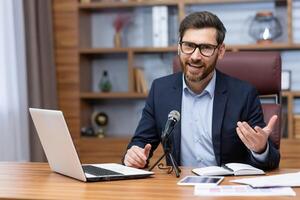 Portrait of successful man inside office, businessman with professional microphone recording online training course and audio podcasts, using laptop sitting at desk working, looking at camera. photo