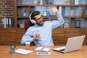 Mature boss investor at workplace inside home office dancing having fun listening to music in headphones, businessman in shirt resting and happy sitting at table using laptop at work. photo