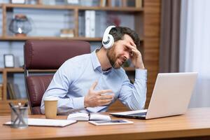 A young male businessman is sitting in the office in headphones and talking on a call from a laptop. Unpleasant conversation, problems with communication, conference. Worriedly holding his head. photo