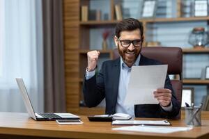 Successful businessman doing paperwork, boss with beard and glasses sitting at desk at workplace using laptop working with documents, holding hand up celebrating victory and triumph. photo